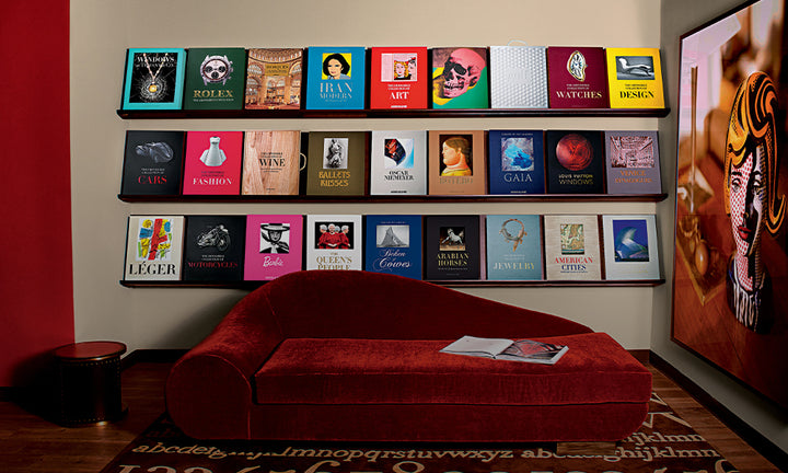 Three shelves of colorful books against a beige wall with a red velvet couch in front